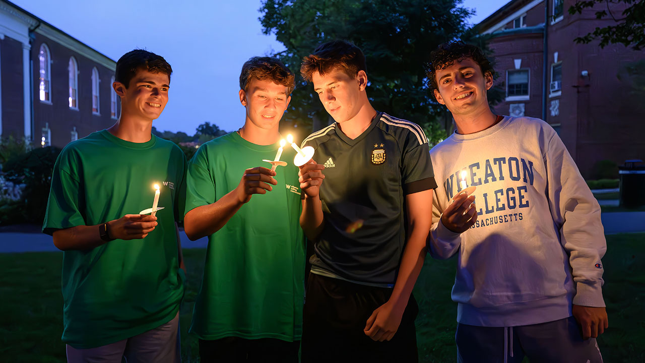 Four male students holding candles outside at night on Chapel Night.